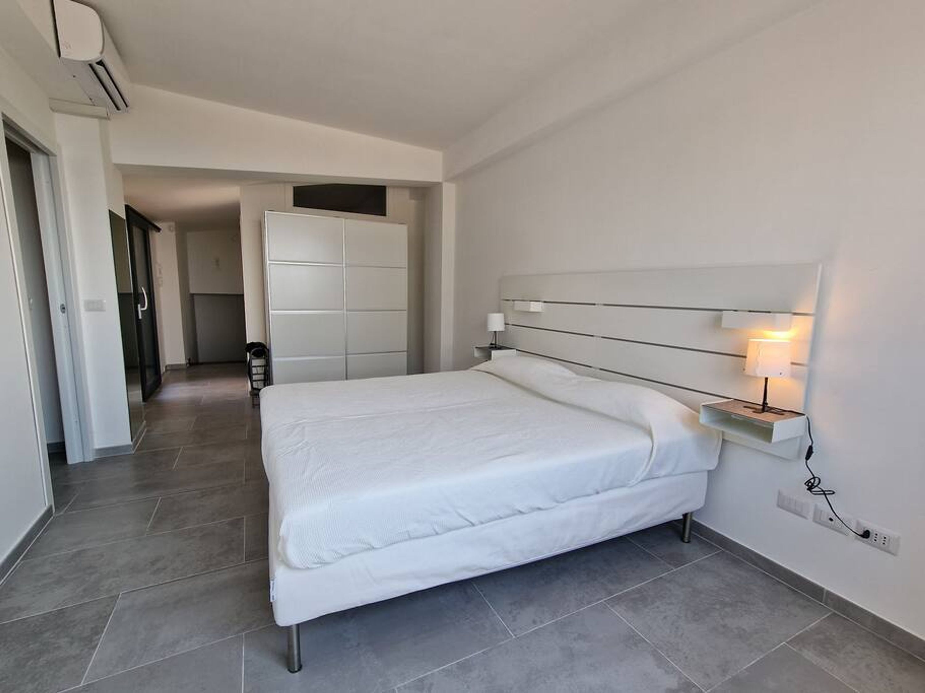 A modern hotel room with a large bed, a bedside table with a lamp, and a wardrobe.