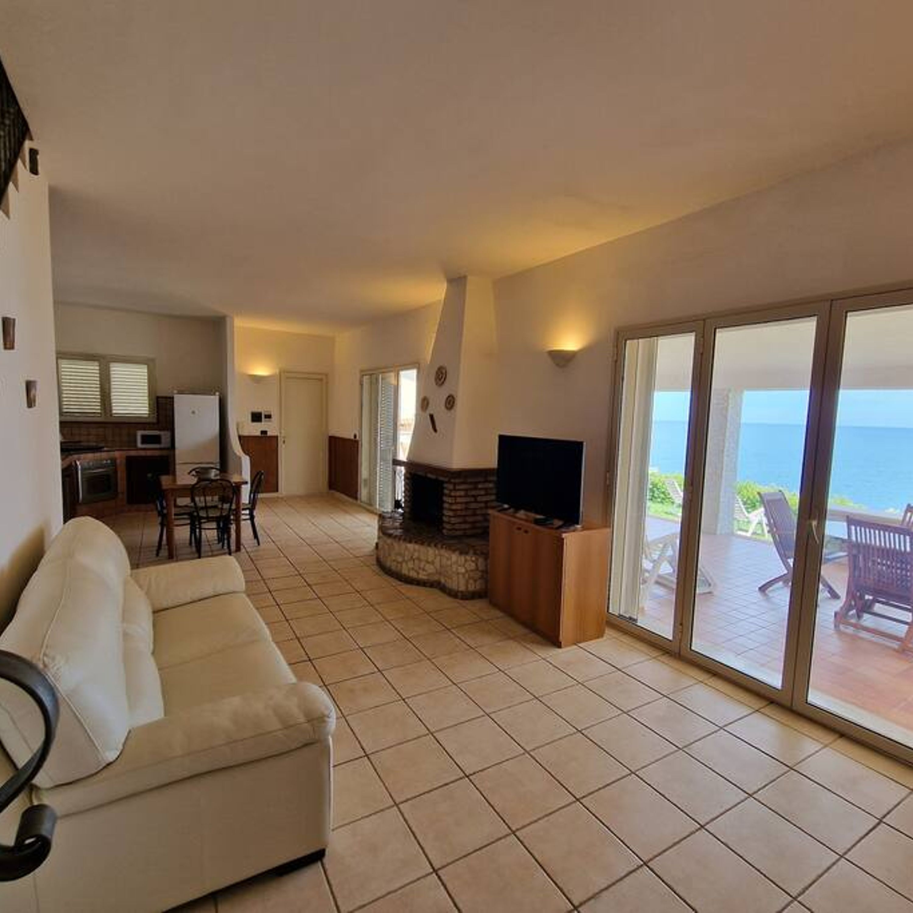 A spacious living area with a fireplace, a sofa, a television, and a balcony with a view of the sea.
