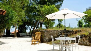 A patio area with a table and chairs under a white umbrella, surrounded by trees and a stone wall. - Villa Bonaccorso - Il Casotto Bouganville (Viagrande)