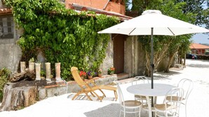 An outdoor seating area with a round table and four chairs, an umbrella, and a wooden chair with a cushion. - Villa Bonaccorso - Il Casotto Bouganville (Viagrande)