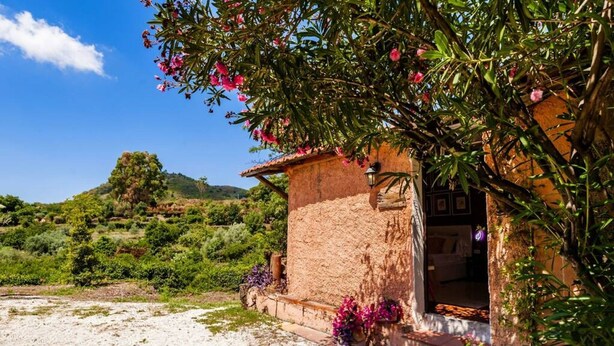 A rustic stone house with a small porch, surrounded by lush greenery and a hill in the background. - Villa Bonaccorso - Il Casotto Bouganville (Viagrande)