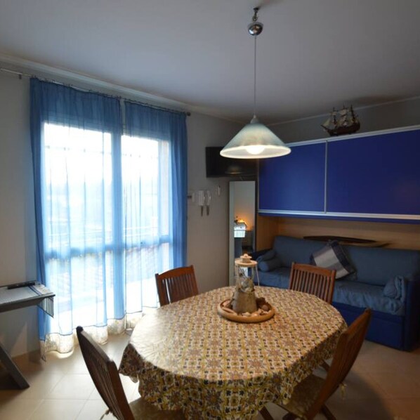 A dining area with a round table, chairs, a hanging light fixture, and a large window with curtains. - Appartamento Orchidea in Residence con Piscina Condominiale (Mascali)