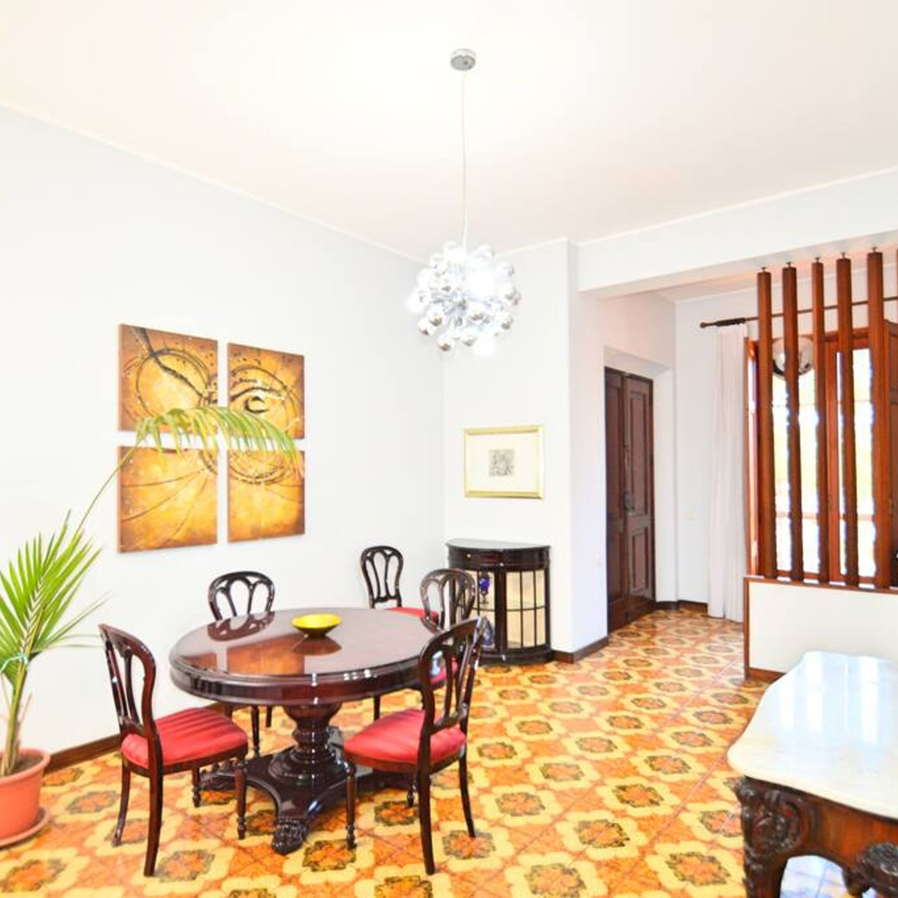 A dining area with a round table, chairs, and a chandelier. There are wall art pieces and a potted plant.