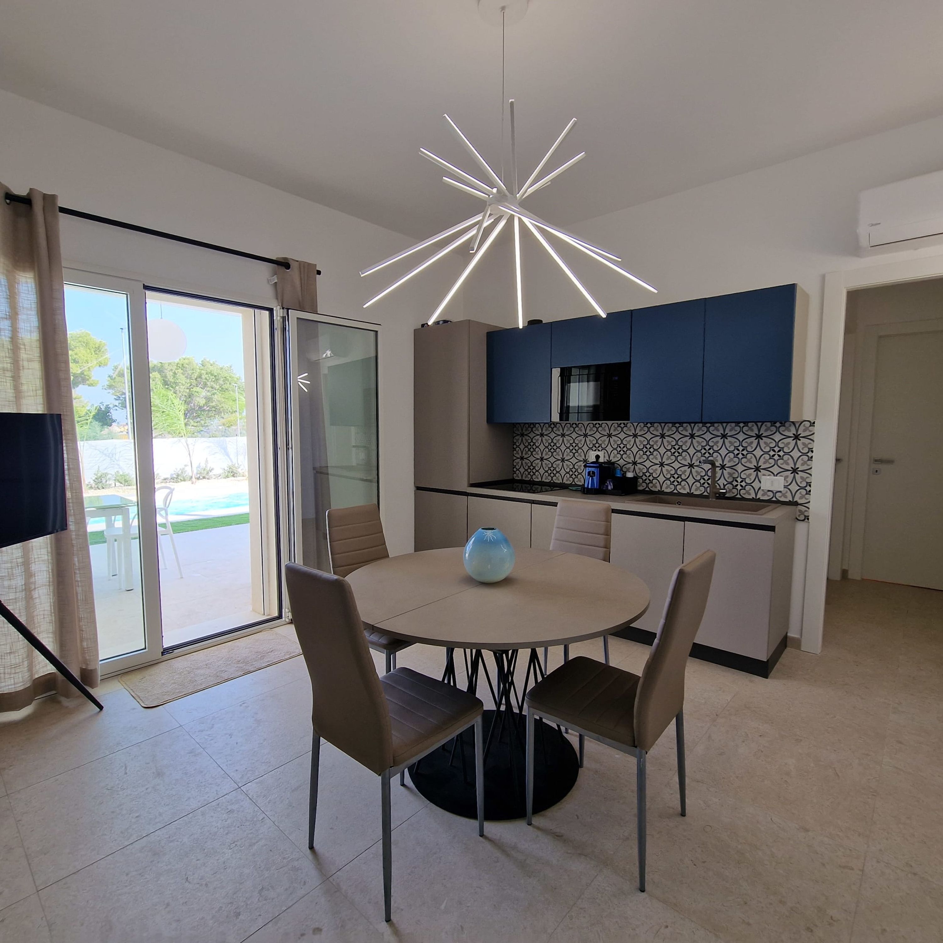 A modern kitchen with a central island, a dining area with four chairs, and a large window with a view of greenery.