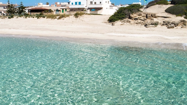Une propriété située en bord de mer, avec une eau turquoise cristalline, un bâtiment blanc aux fenêtres bleues et une plage de sable.