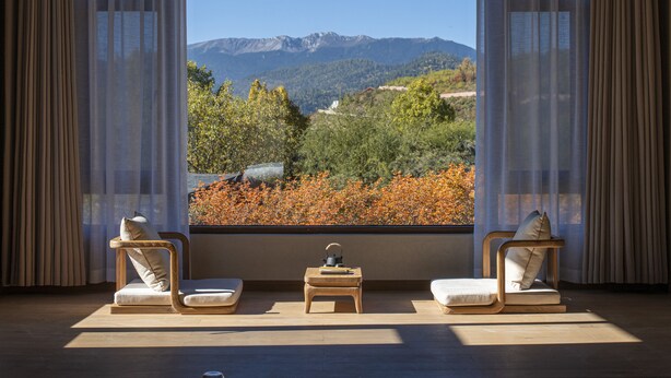 A room with a large window offering a mountain view, two wooden chairs, a small table, and sheer curtains. - Shujia wengmacuozangshidujiazhuangyuan (Deqin)