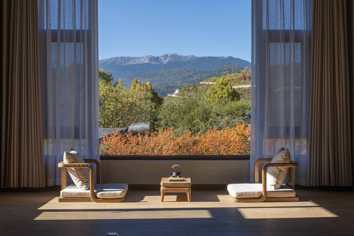 Une pièce dotée d’une grande fenêtre donnant sur les montagnes, de deux chaises en bois, d’une petite table et de rideaux légers.