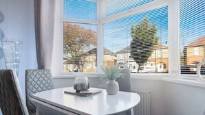 Un coin repas avec une table blanche, des chaises grises à revêtement en tissu, un vase avec une plante et une fenêtre avec des stores.