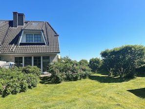A residential house with a sloped roof, a chimney, and a well-maintained lawn with flowering bushes.