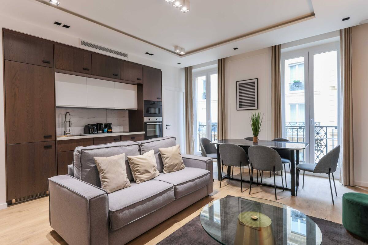 A modern living room with a grey sofa, a glass coffee table, and a dining area with a table and chairs.