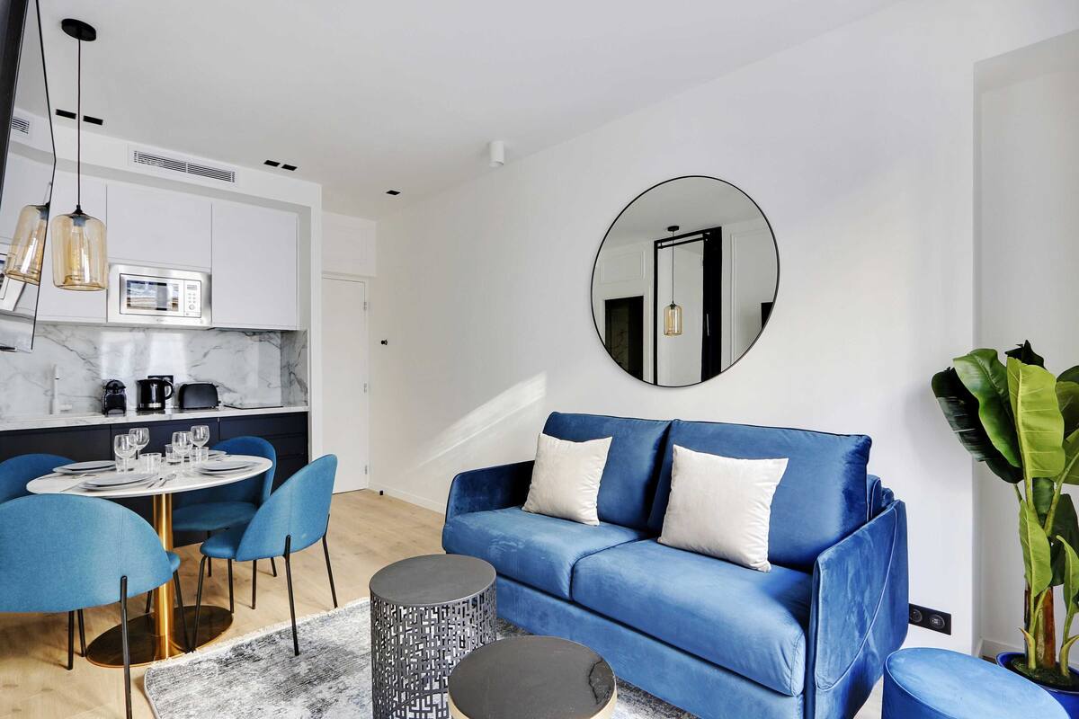 A modern living room with a blue sofa, round coffee table, and matching chairs. A kitchen with white cabinets and a microwave is visible in the background.
