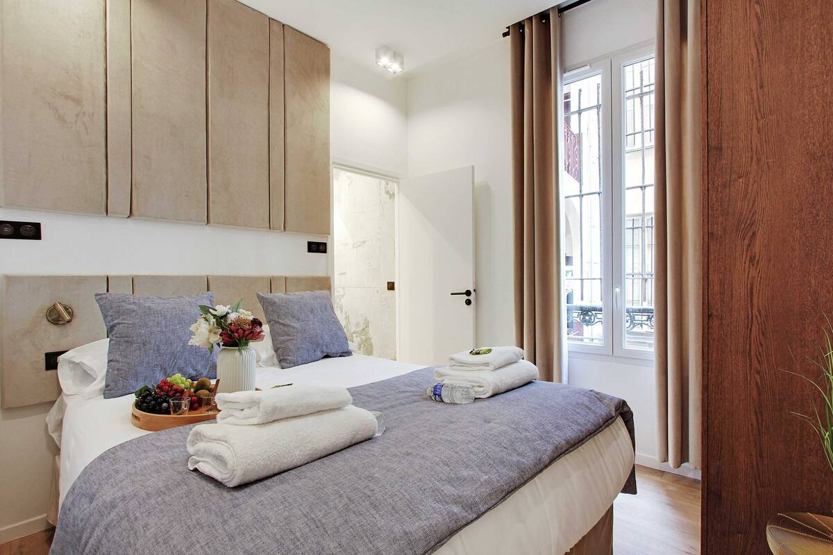 A hotel room with a bed, a wooden headboard, a window with curtains, and a small table with towels and fruit.