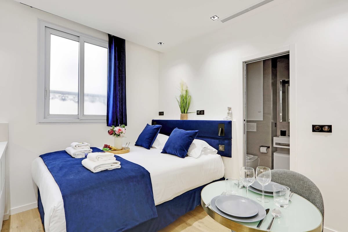 A modern hotel room with a large bed, a dining table with plates and glasses, and a view of the outdoors through a window.