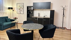 Een moderne woonkamer met een groene bank, een ronde tafel met stoelen, een keukenhoek met donkere kasten, een magnetron en een vloerlamp.