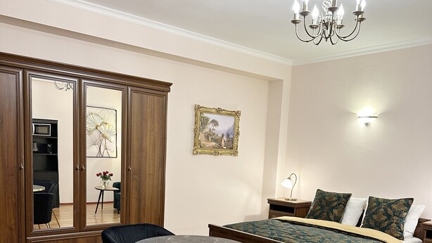 Een moderne woonkamer met een ronde tafel, twee zwarte stoelen, een bank, een bed met beddengoed met een patroon, een kroonluchter en een ingelijst schilderij aan de muur.