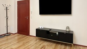 Een kamer met een houten vloer, een kapstok, een rode deur, een zwarte entertainmentkast met een flatscreen televisie erboven, en een klein decoratief voorwerp op de kast.