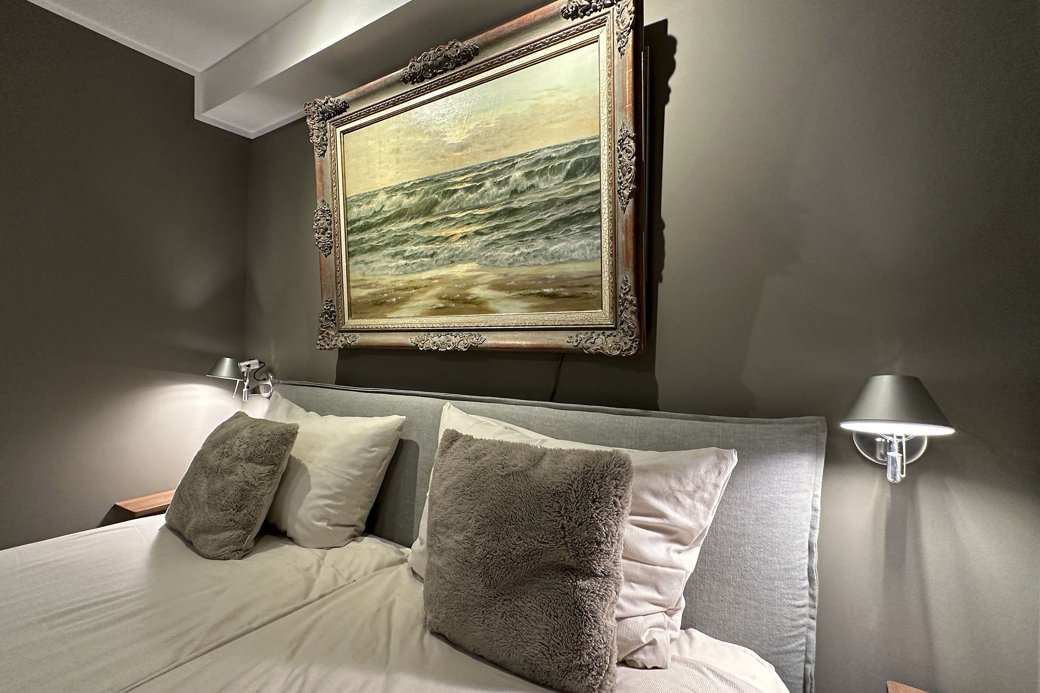 Une chambre à coucher avec un grand lit, deux oreillers, une table de chevet, une lampe fixée au mur et un tableau encadré représentant un paysage marin.