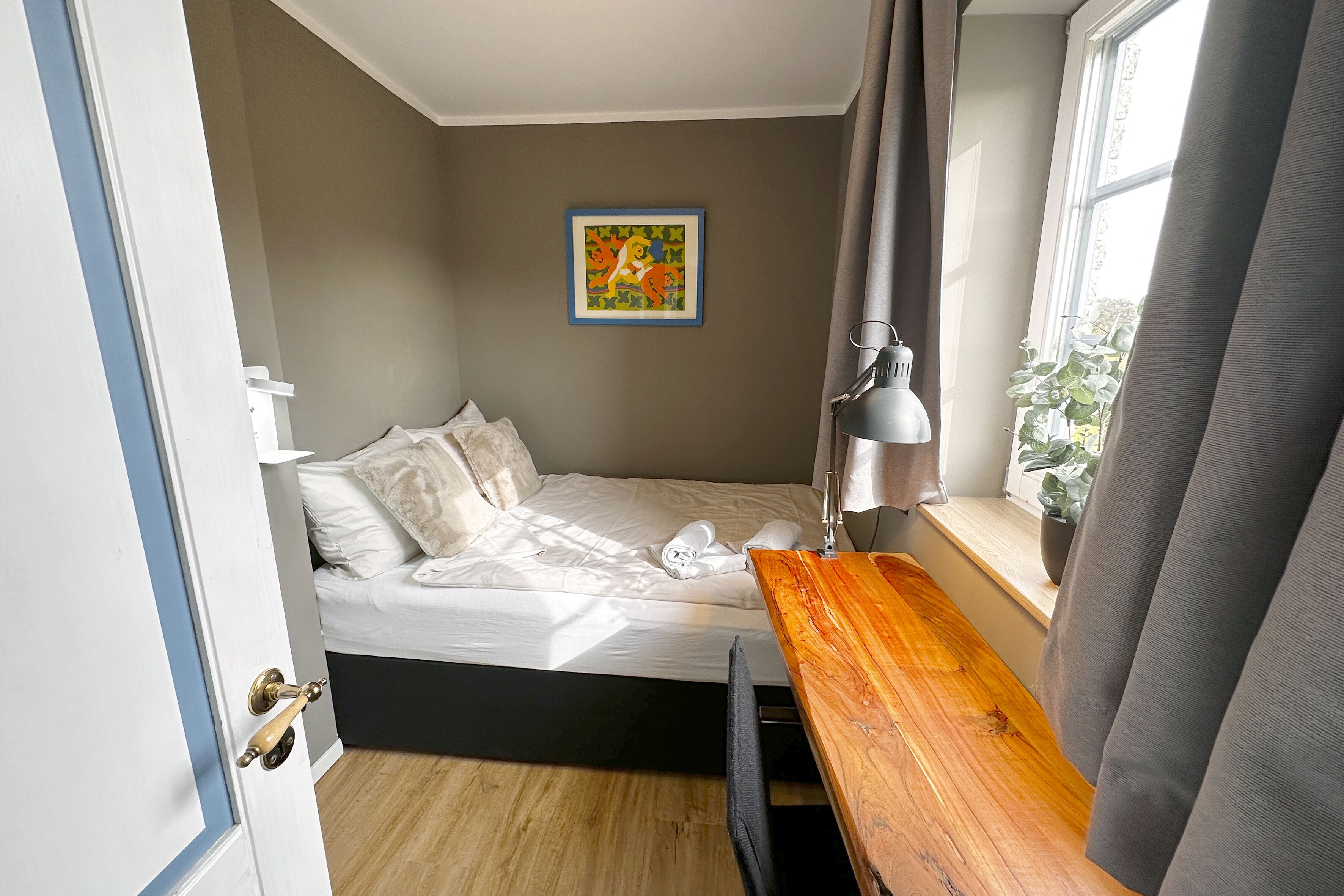 A bedroom with a bed, a bedside table, a window, and a painting on the wall.