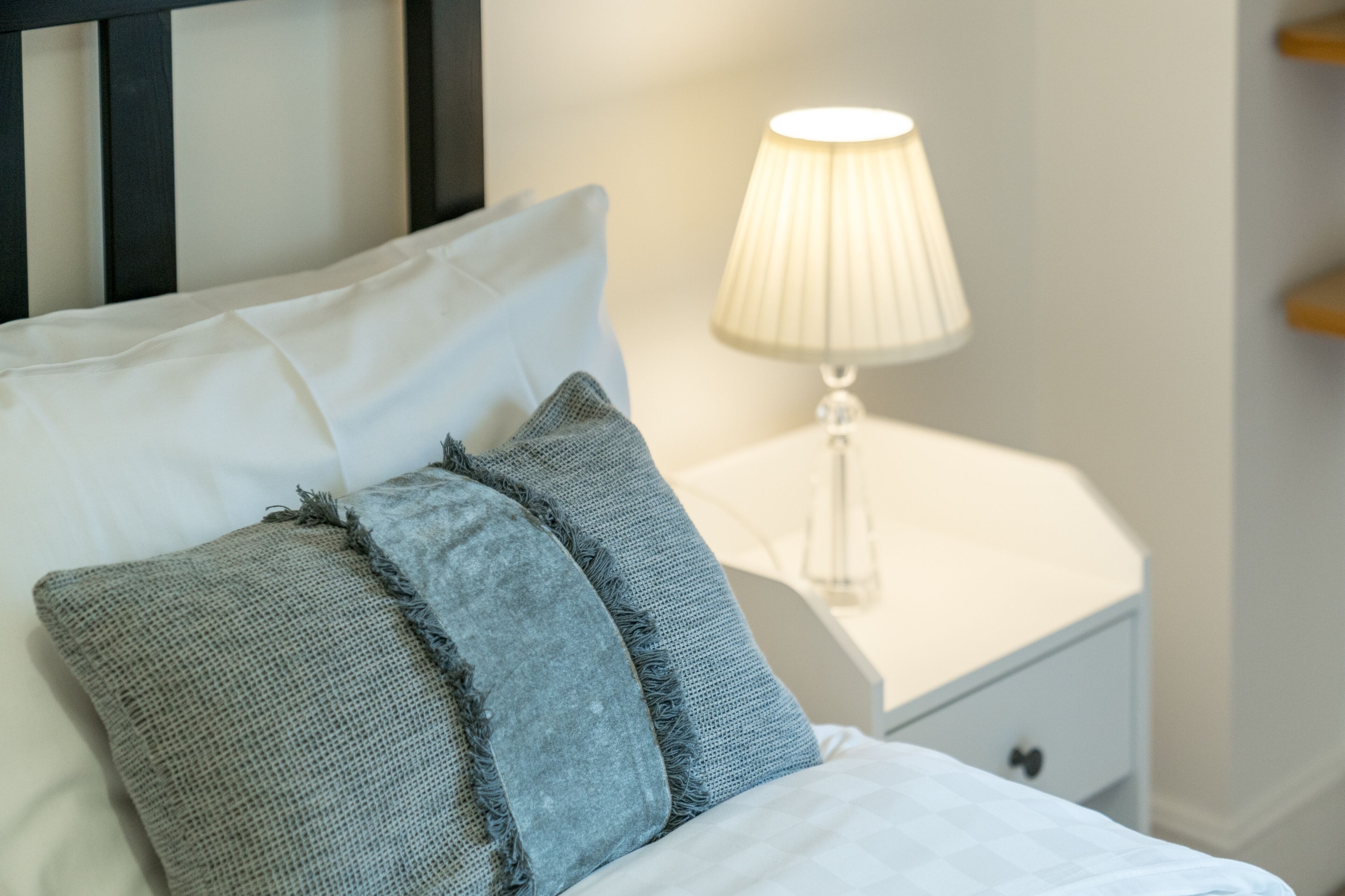 En pænt redt seng med hvide og grå puder, et hvidt natbord med en lampe og en lille skuffe.