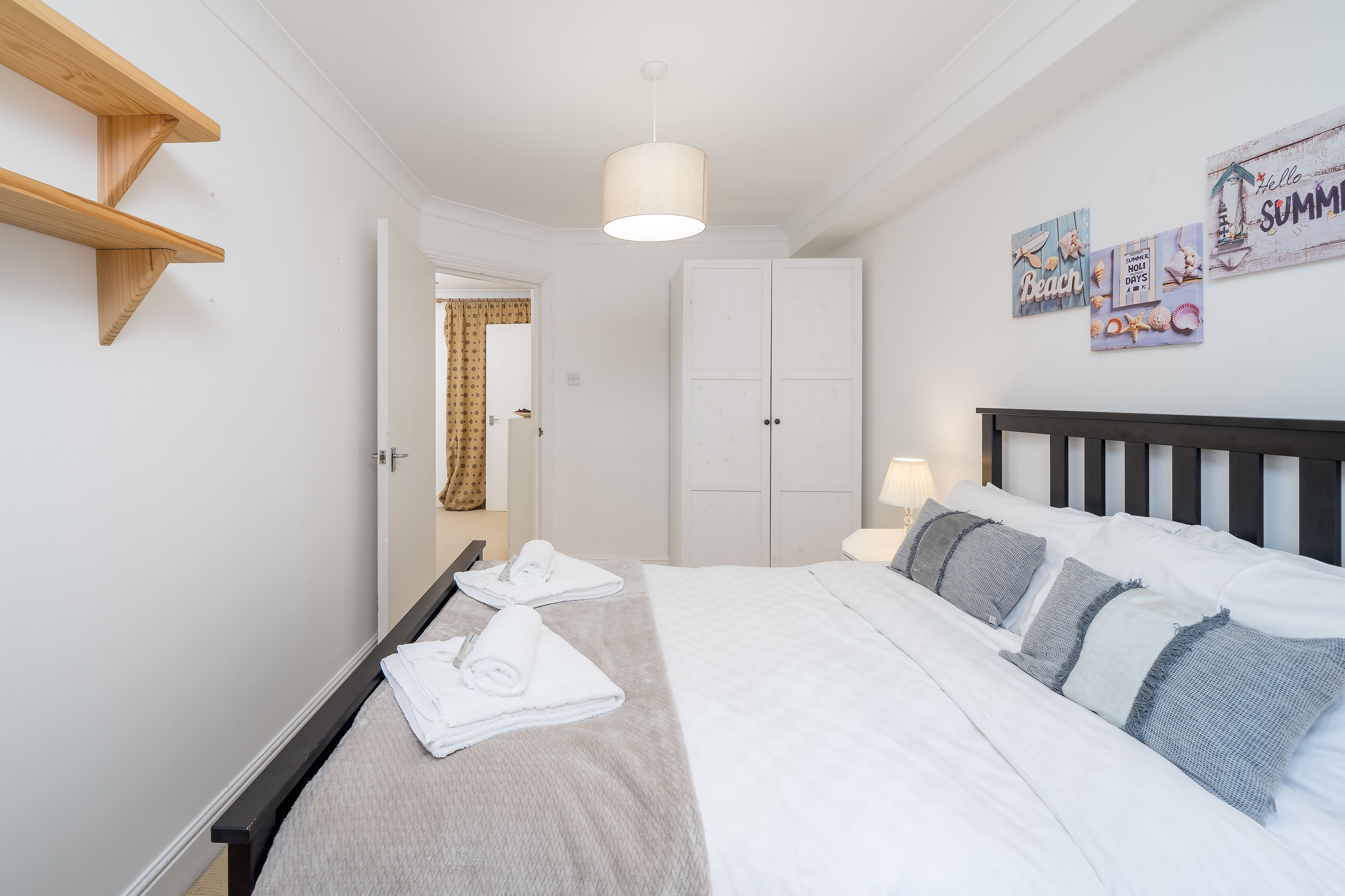 A neatly made bed with white linens and rolled towels, a wooden headboard, and a wall with a 'Hello Summer' sign.