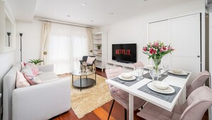 En moderne stue med sofa, sofabord og spisebord dækket til to personer. Et fladskærms-tv er monteret på væggen, og der står en vase med blomster på spisebordet.