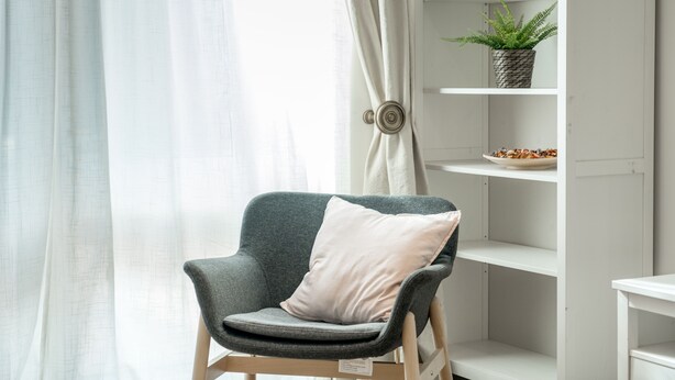 En moderne stue med en grå lænestol, en hvid reol med en plante, og et vindue med lette gardiner.