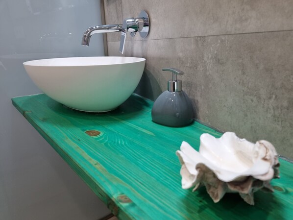 A bathroom with a white vessel sink, a green wooden shelf, a soap dispenser, and a seashell.
