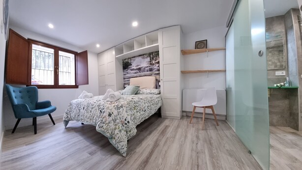 Un dormitorio con cama, una silla azul, una silla blanca, un armario y un área de ducha.