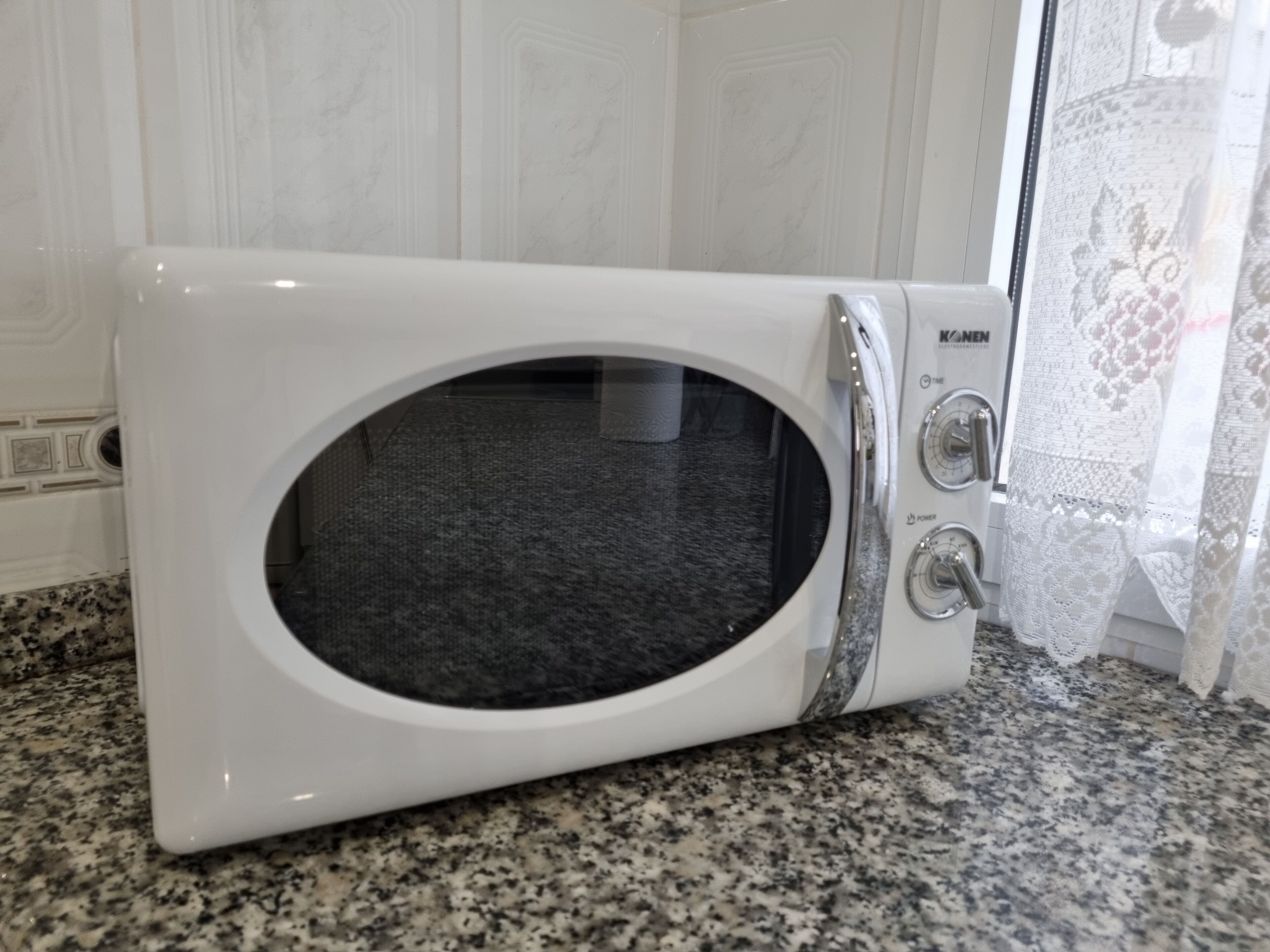 Un four à micro-ondes blanc est posé sur un plan de travail en granit dans une cuisine aux meubles blancs et ornée d’un rideau en dentelle.