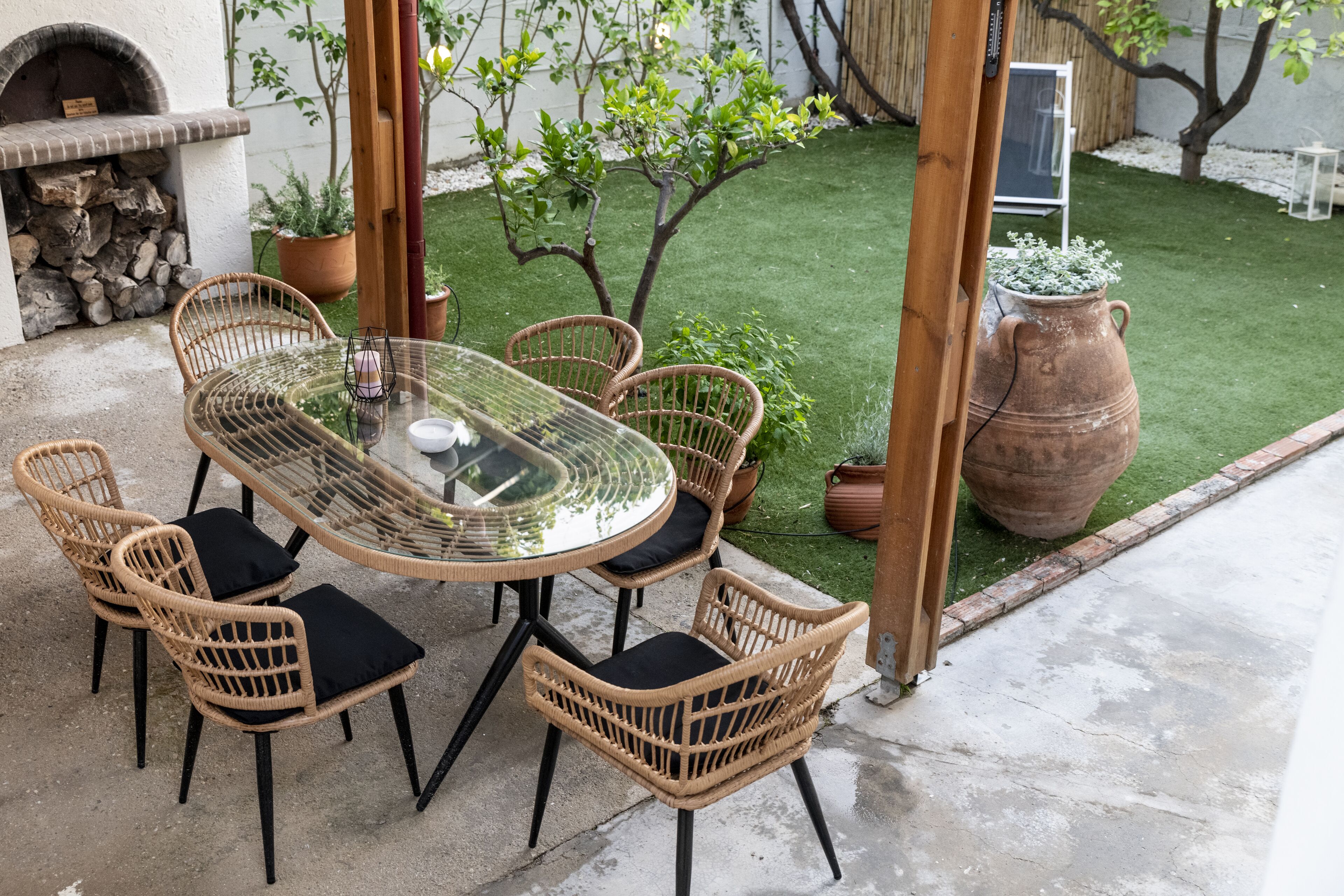 A glass-top table with wicker chairs and black cushions, surrounded by potted plants and a small tree.