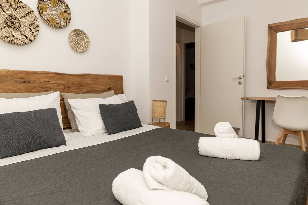 A neatly made bed with grey bedding and white towels, a wooden headboard, and decorative woven wall art.
