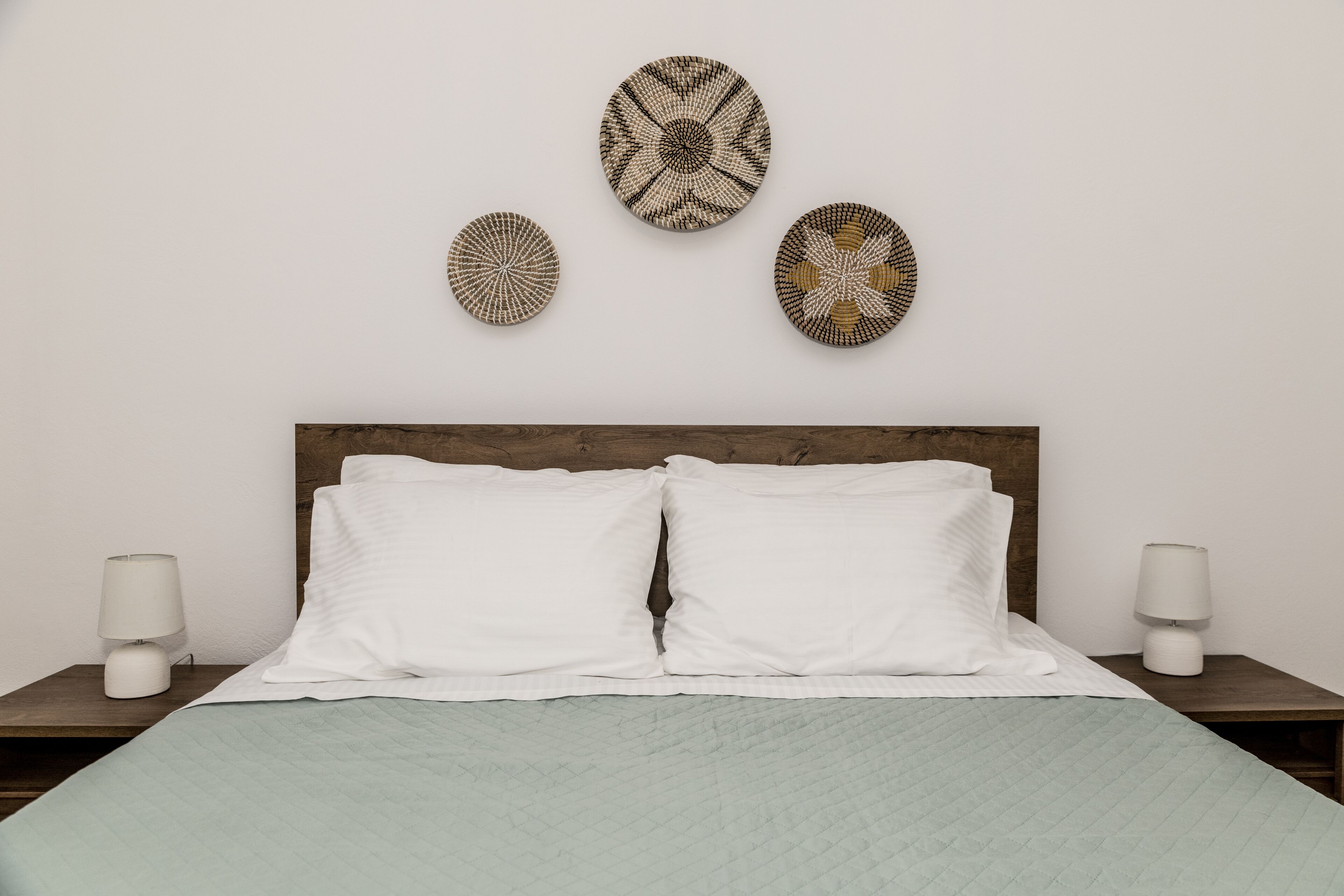 A neatly made bed with white pillows and a light green quilt, flanked by two bedside tables with white lamps, and three wall-mounted decorative plates above the headboard.