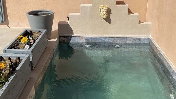 Una piscina con una fuente de piedra, una ventana con contraventanas azules y una jardinera con flores.