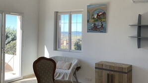 Una habitación con techo de madera, un ventanal grande, una silla, un cuadro y un armario pequeño.