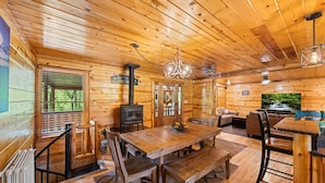 A wooden cabin interior with a dining area, a fireplace, a television, and a large window overlooking greenery. - Tequila Moon: Dive Into Paradise (Sevierville)