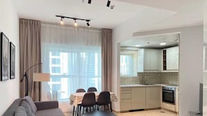 Ein modernes Wohnzimmer mit Sofa, Esstisch und Stühlen, Küchenzeile mit Schränken und Backofen sowie einem großen Fenster mit Vorhängen.