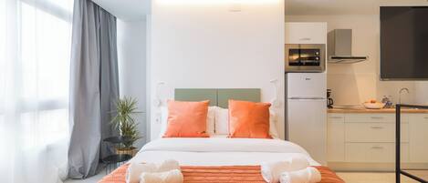 Ett modernt hotellrum med en stor säng, orange sängkläder, ett litet kök och ett stort fönster med gardiner.