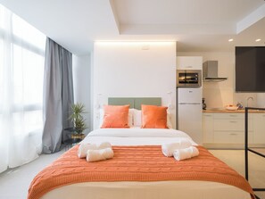 A modern hotel room with a large bed, orange bedding, a kitchenette, and a large window with curtains. - Shabolo Apartments las Palmas (Las Palmas de Gran Canaria)