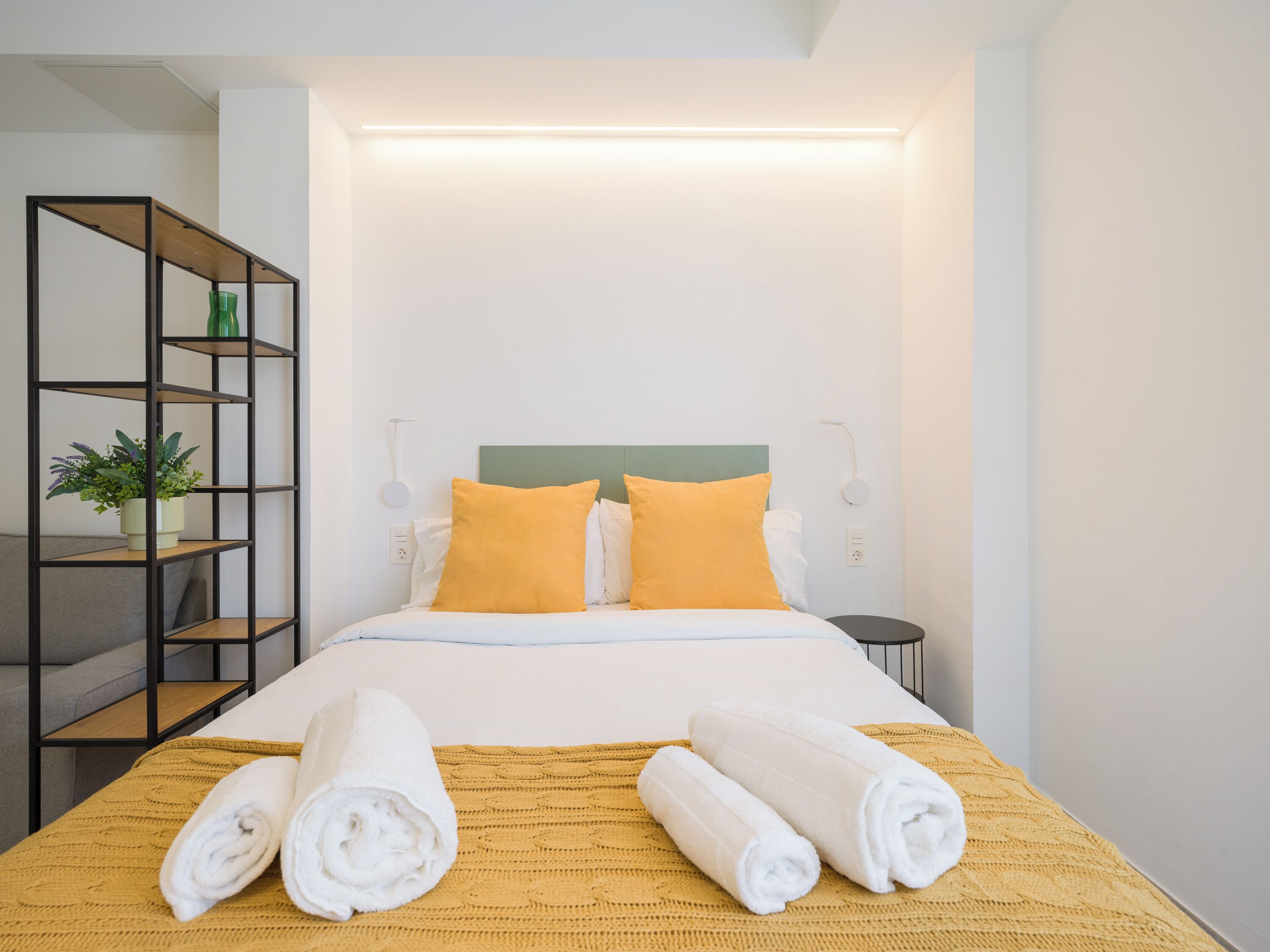 A neatly made bed with two pillows, a bedside table, a lamp, and a shelf with a plant.