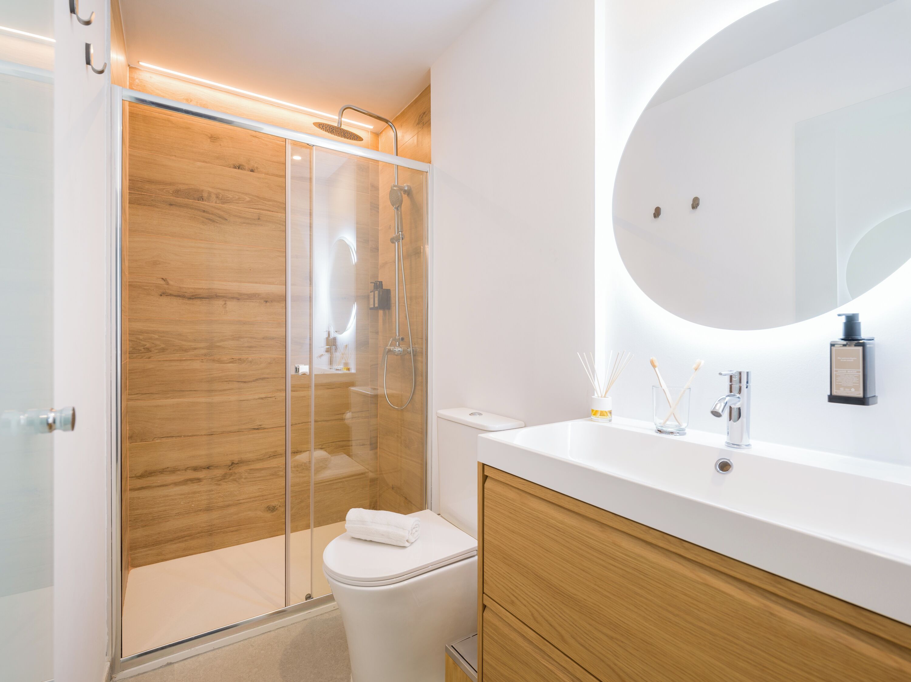 Une salle de bain moderne avec une douche à parois de verre, des toilettes blanches, un meuble-lavabo en bois et un grand miroir.