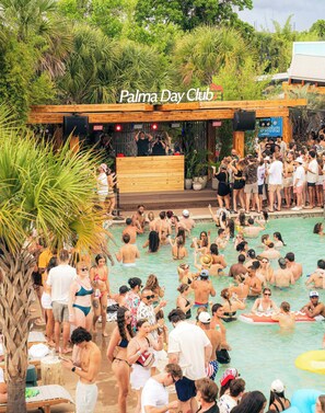 A crowded outdoor pool area with a wooden deck and a stage for live music.