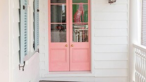 A porch with a pink door, white siding, and a wooden floor. - Casa Zoë | Royal Cottage I – Designer Escape (Charleston)