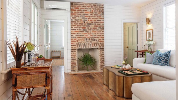 A cozy living room with a brick fireplace, white walls, and a white sofa. - Casa Zoë | Royal Cottage I – Designer Escape (Charleston)