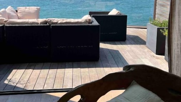 Un balcón con un sofá negro y una silla de madera, con vista a un cuerpo de agua y terreno distante.