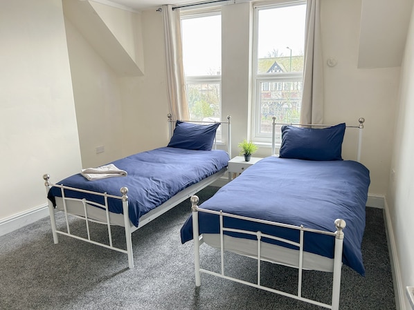 A bedroom with two single beds, a small bedside table with a plant, and a window with a view of buildings.