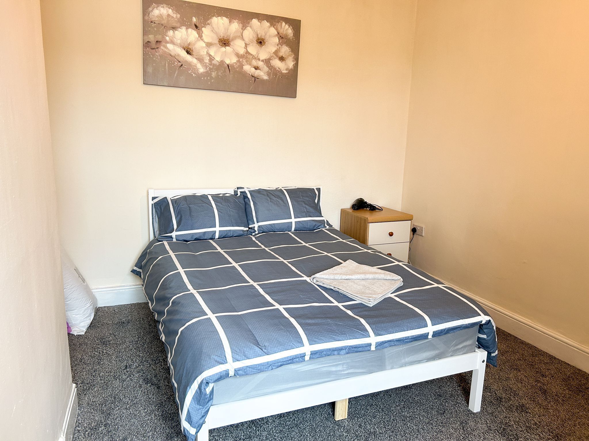 A neatly made bed with a blue and white checkered bedspread, a wooden nightstand with a lamp, and a painting of flowers on the wall.