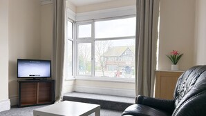 Una sala de estar con un sofá de cuero negro, una mesa de centro blanca, un televisor sobre un mueble de madera y una ventana con cortinas.