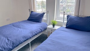 Una habitación pequeña y luminosa con dos camas individuales, una mesita con una planta y una ventana que ofrece vistas a edificios.