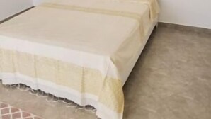 A single bed with a beige bedspread in a room with white walls and a tiled floor.
