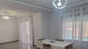 A spacious dining area with a table and chairs, a ceiling light fixture, and large windows with curtains.